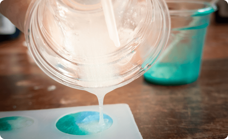Pouring white resin over a blue and green coaster on a wooden surface.